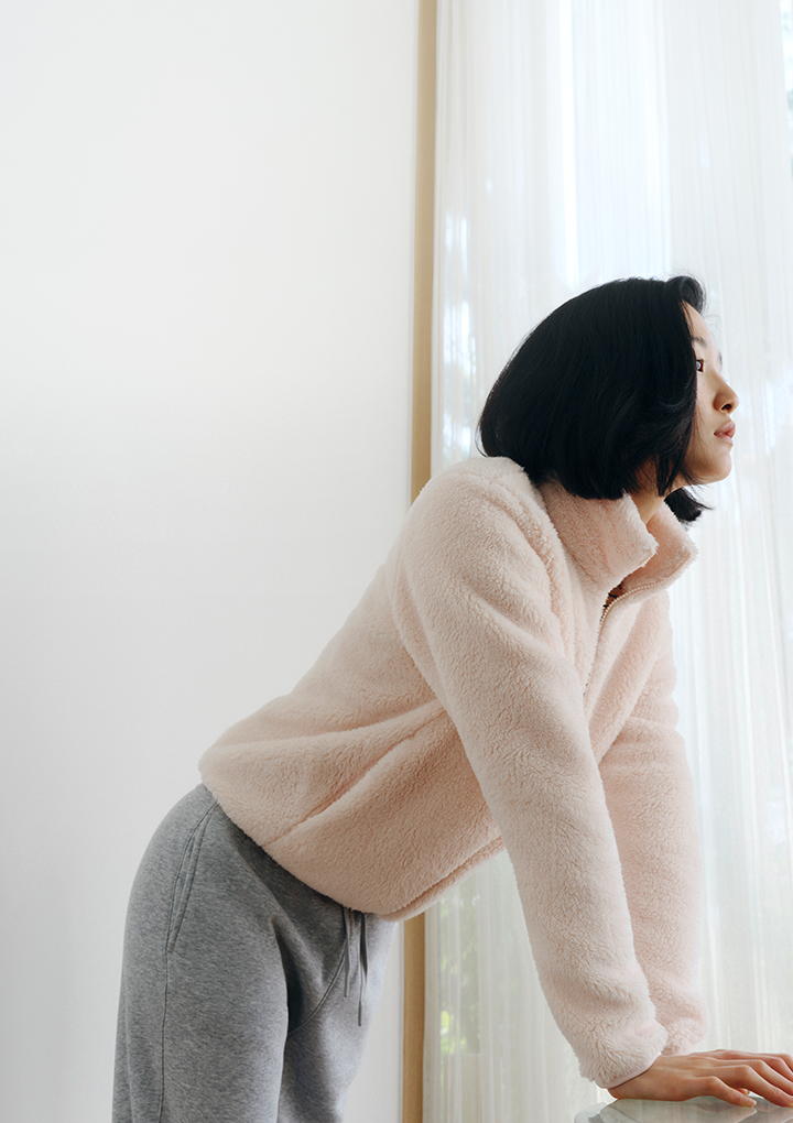 창가에서 핑크 플러피 플리스 집업과 그레이 조거팬츠를 입고 기대어 선 여성 모델